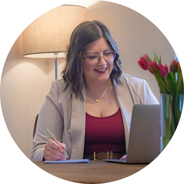 arbeiten-am-laptop Jenny Schmitz sitzt an ihrem Schreibtisch, in einem Raum mit den Farben beige, salbei-grün und beere. Sie sitzt am Laptop und schaut lächelnd nach unten