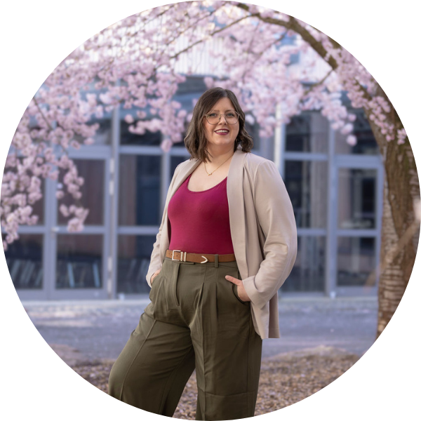 Jenny Schmitz steht unter einem Dach von Kirschblüten. Sie hat die Hände smart in den Taschen ihrer grünen Stoffhose und lächelt in die Kamera